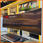 A curved all-in-one computer monitor displaying a boot screen with the AMI logo and terminal text, set on a desk with a keyboard, mouse, and laptop in a retail environment.